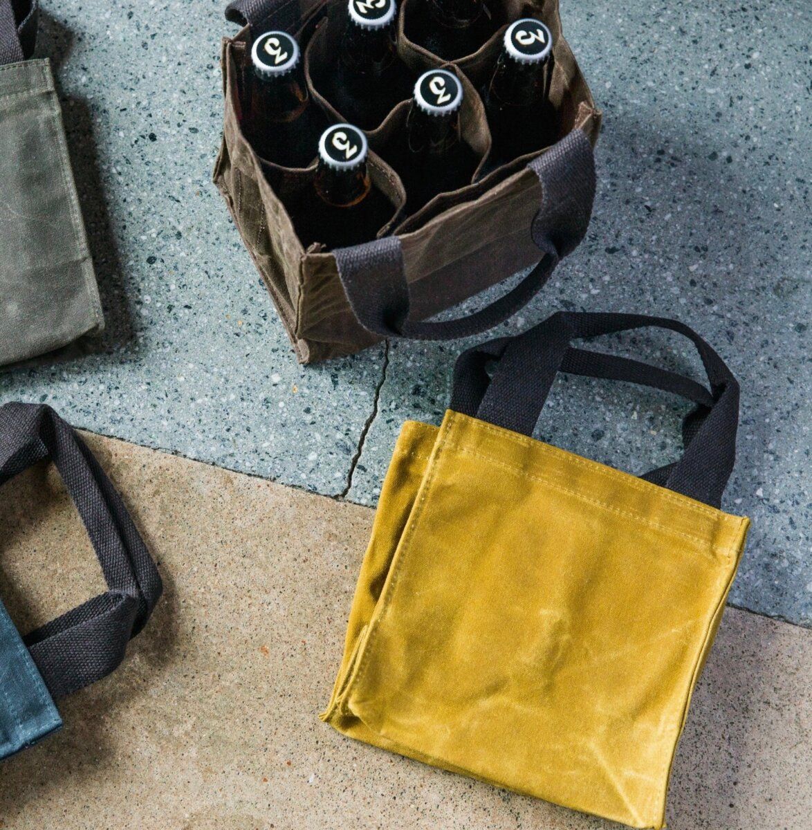 Four waxed canvas tote bags are on a concrete floor; one bag holds six capped glass bottles, while the others—part of our August FINDS—are empty and displayed in vibrant yellow, blue, and green colors, perfect for adding a pop of Nashville style to your daily routine.