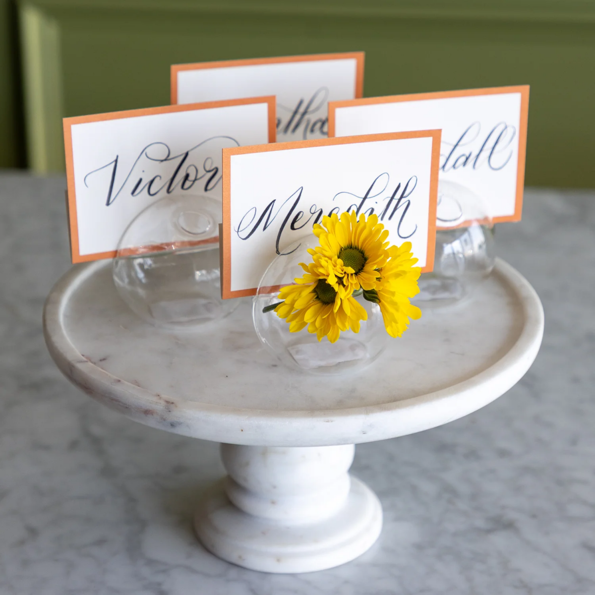 Four handwritten place cards in glass holders are displayed on a marble stand, with one holder containing a small bunch of yellow flowers—a charming tablescape perfect for August FINDS in Nashville.