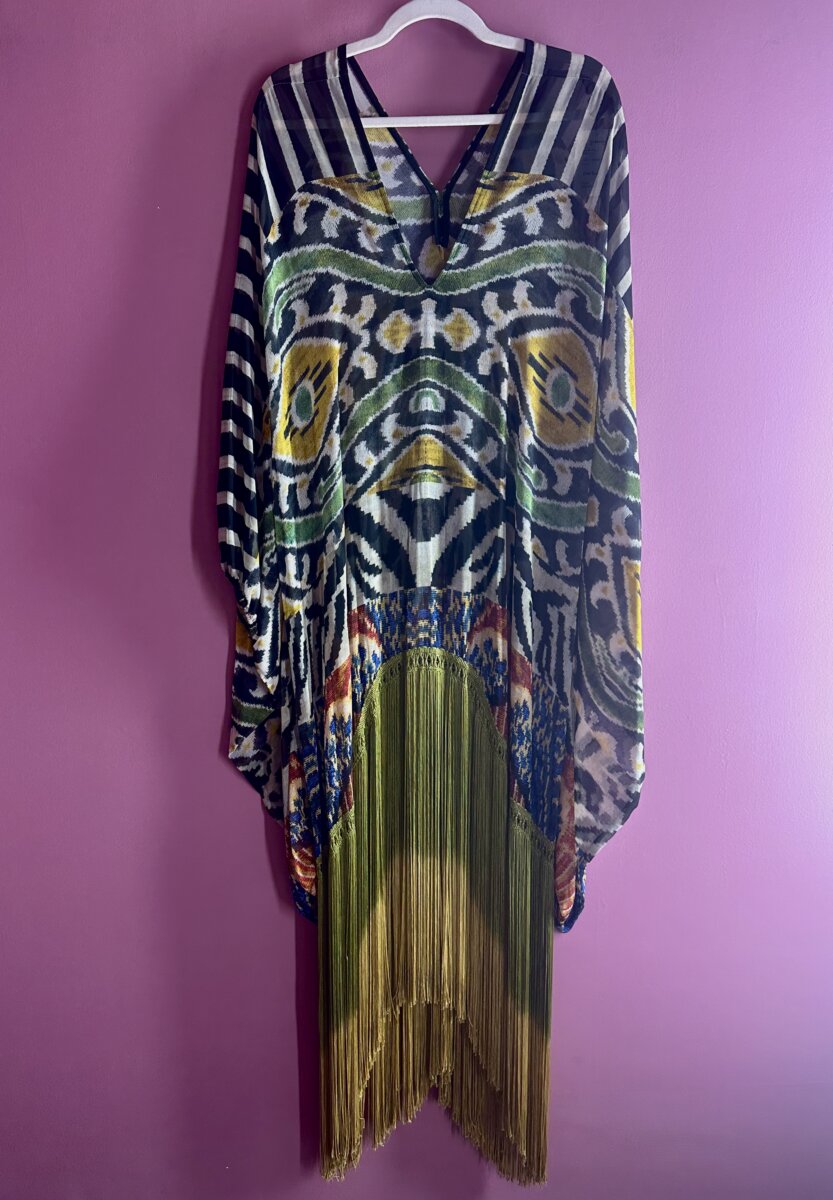 A colorful caftan with fringe, hanging up on a purple wall.