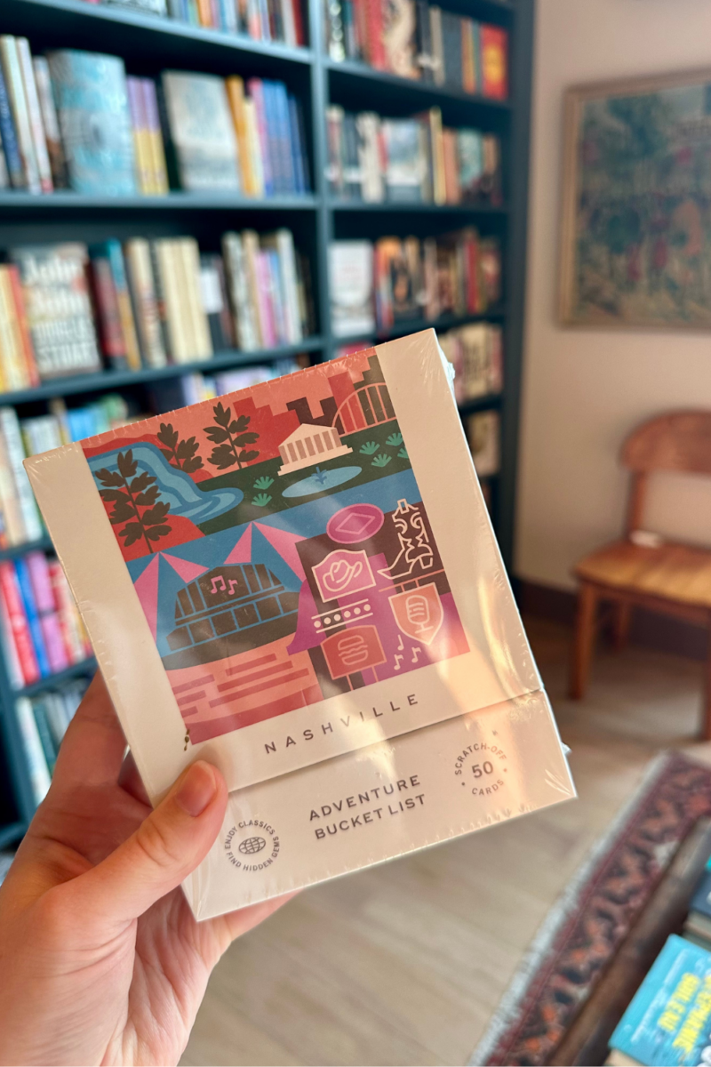 A hand holds a boxed set labeled “Nashville Adventure Bucket List” in a bookstore, with shelves of books and a wooden chair visible in the background—a perfect August FINDS pick for Nashville explorers.