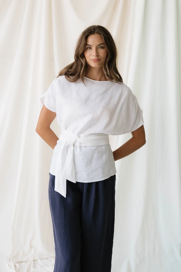 Woman stands in front of white drapes wearing a loose white blouse with a tie belt and dark wide-leg pants, hands behind her back.