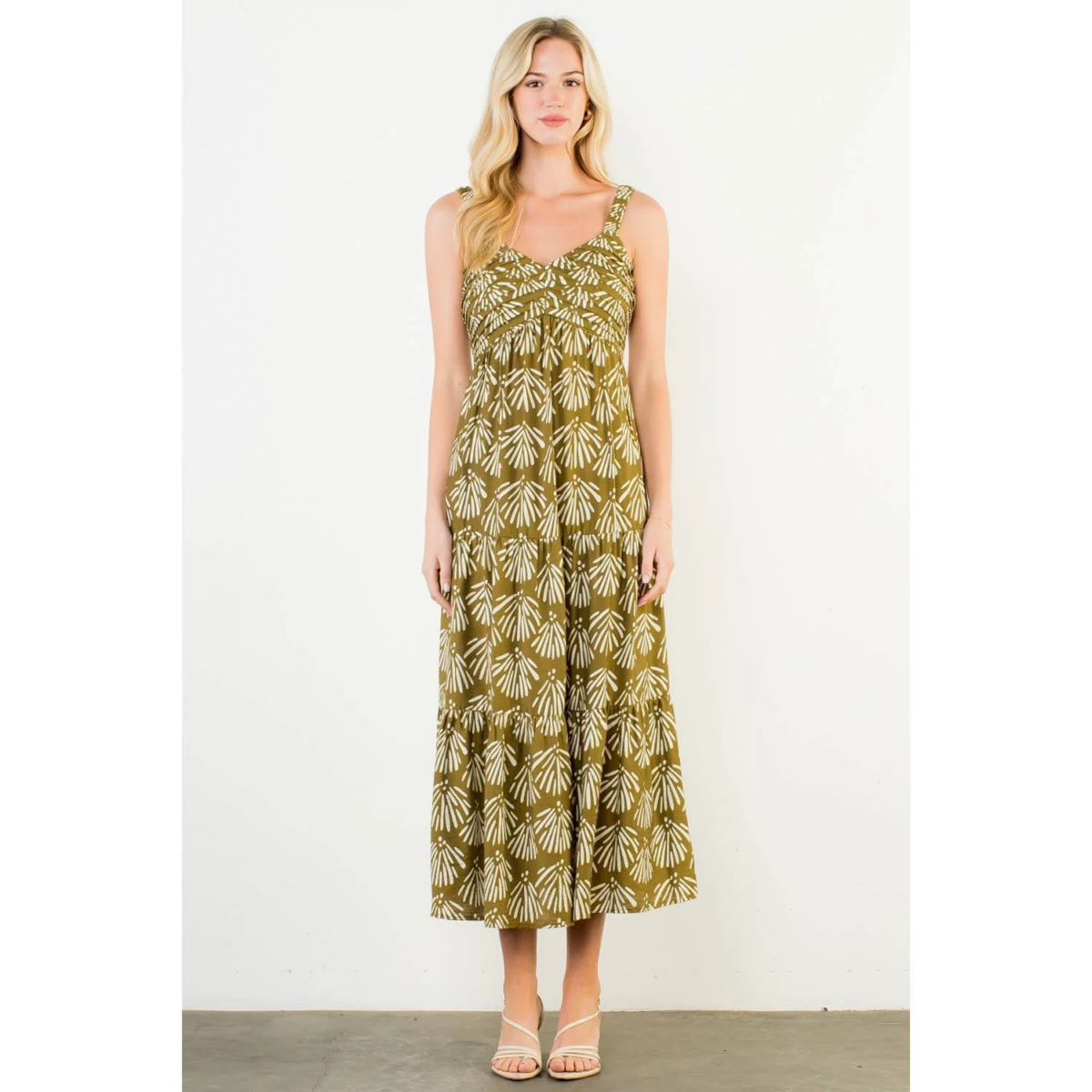 Woman standing against a white wall wearing a long, sleeveless olive-green dress with a white leaf pattern and beige sandals—a perfect look for those August FINDS in Nashville.