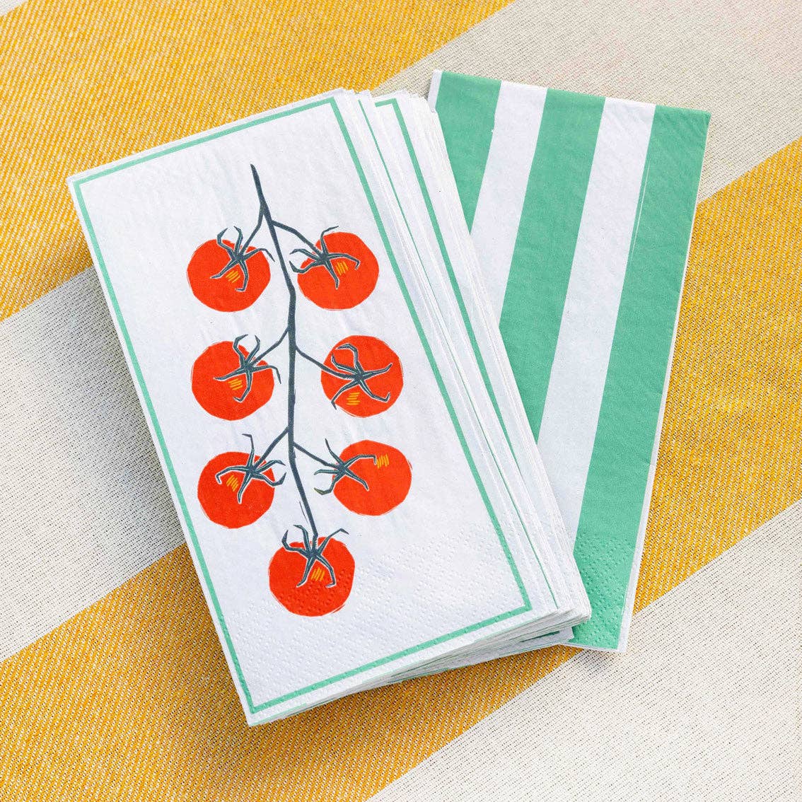 A stack of napkins, some with a tomato vine illustration and some with green and white stripes, placed on a yellow and white striped surface.