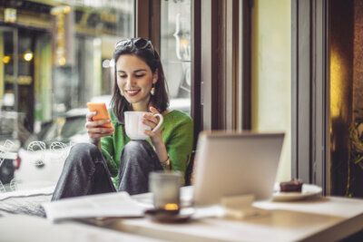 Woman sitting by a window in a cafe, holding a mug and looking at her phone, with a laptop and papers on the table in front of her.