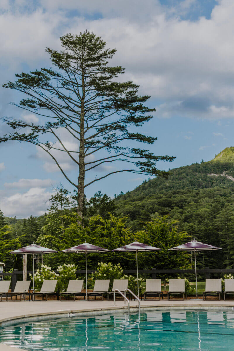 8 Luxurious, Family-Friendly Resorts in the South - 4 Lounge chairs and closed umbrellas line the edge of a clear outdoor pool at one of the region’s top Family-Friendly Resorts, with tall trees and green hills in the background under a partly cloudy sky.