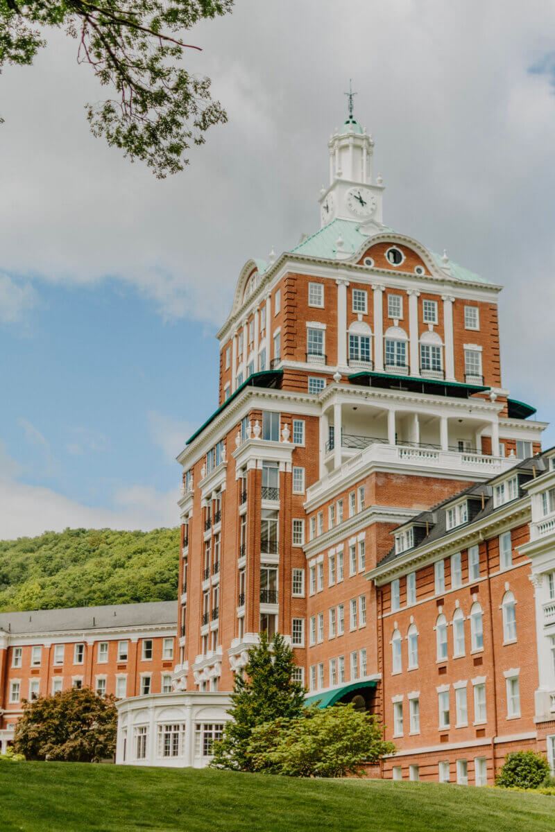 8 Luxurious, Family-Friendly Resorts in the South - 2 A large historic red-brick building with white columns and a clock tower, surrounded by trees and hills under a partly cloudy sky, evokes the charm found at family-friendly resorts popular among Southern families.