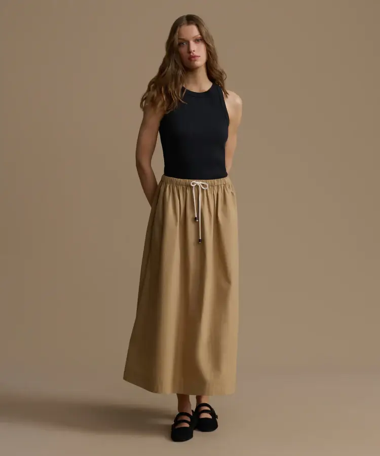 A person stands against a plain background, showcasing Summer Essentials in style with a black sleeveless top, a beige drawstring maxi skirt, and black shoes.