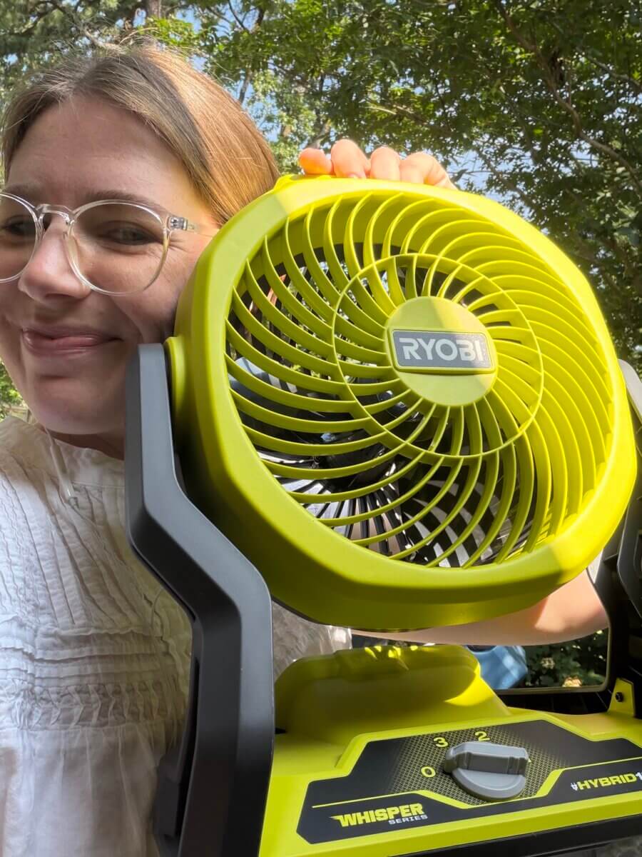 Smiling at the camera, a person holds a bright yellow Ryobi fan outdoors, framed by green trees—capturing a perfect summer moment for the SB Hot List, July 2026.