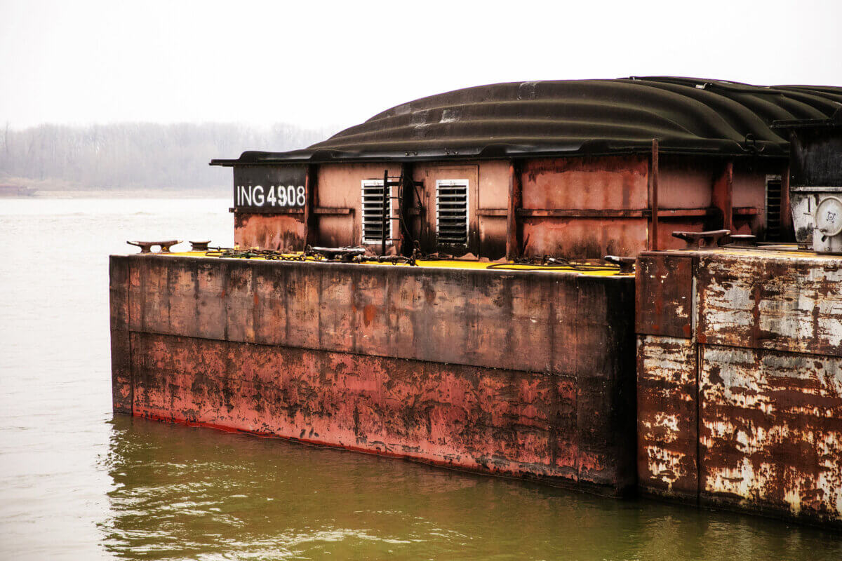 Rusty barge labeled "ING 4908" floats on murky Mississippi water near a shoreline with bare trees in the background under an overcast sky.