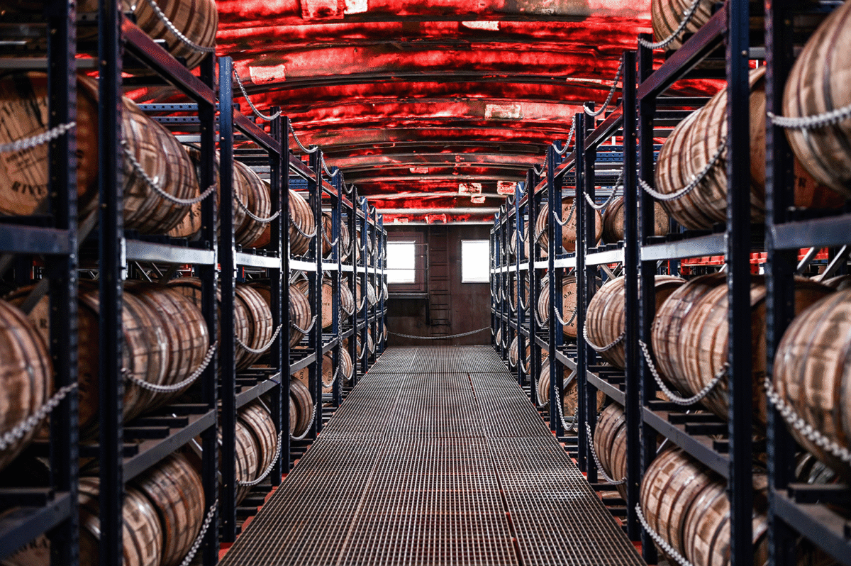 Rows of wooden barrels on metal racks line both sides of a warehouse with a red ceiling and grated metal floor, viewed from the center aisle—a classic scene from a Kentucky distillery renowned for its bourbon craftsmanship.
