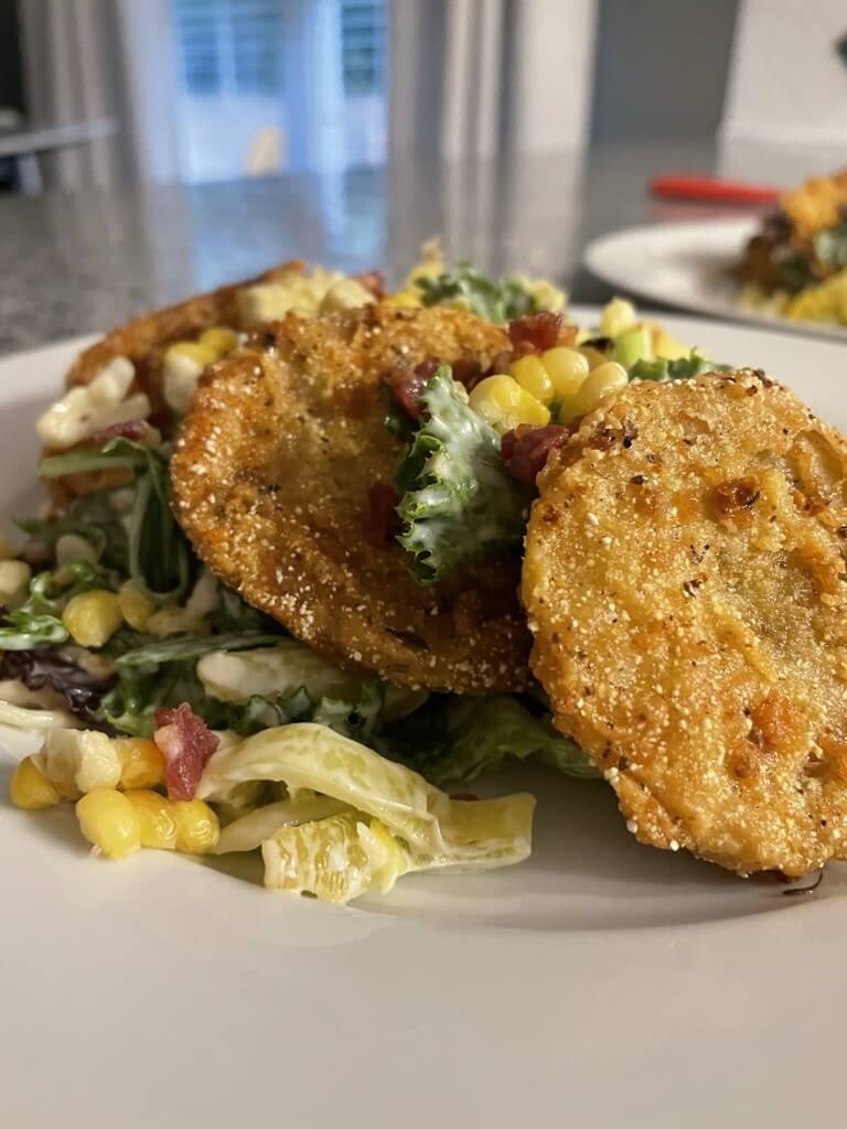 A close-up of a salad topped with fried green tomato slices, corn, and bacon bits on a white plate—bringing a taste of Mississippi comfort to the table, perfect for pairing with a splash of Kentucky bourbon from your favorite distillery.