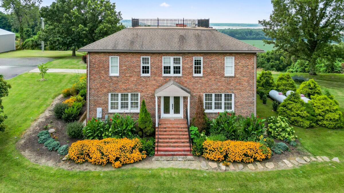 Two-story brick house with a central entrance, white-framed windows, and a landscaped front yard with yellow flowers and green shrubs, set on a grassy lot with trees and open fields in the background.