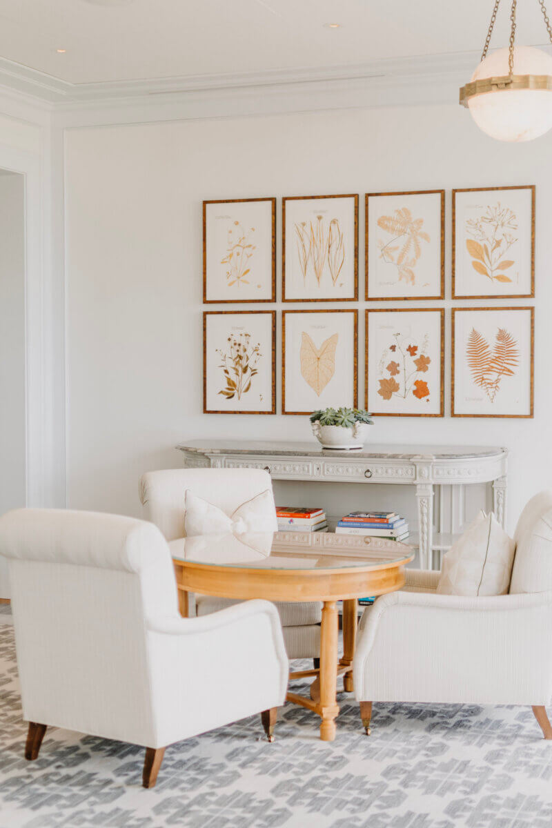 8 Luxurious, Family-Friendly Resorts in the South - 13 Two white armchairs and a round wooden table are in front of a console with a plant, creating a cozy atmosphere reminiscent of the inviting lounges found at family-friendly resorts. Botanical prints in wooden frames are arranged in a grid on the white wall above, striking just the right balance of comfort and style ideal for Southern families.