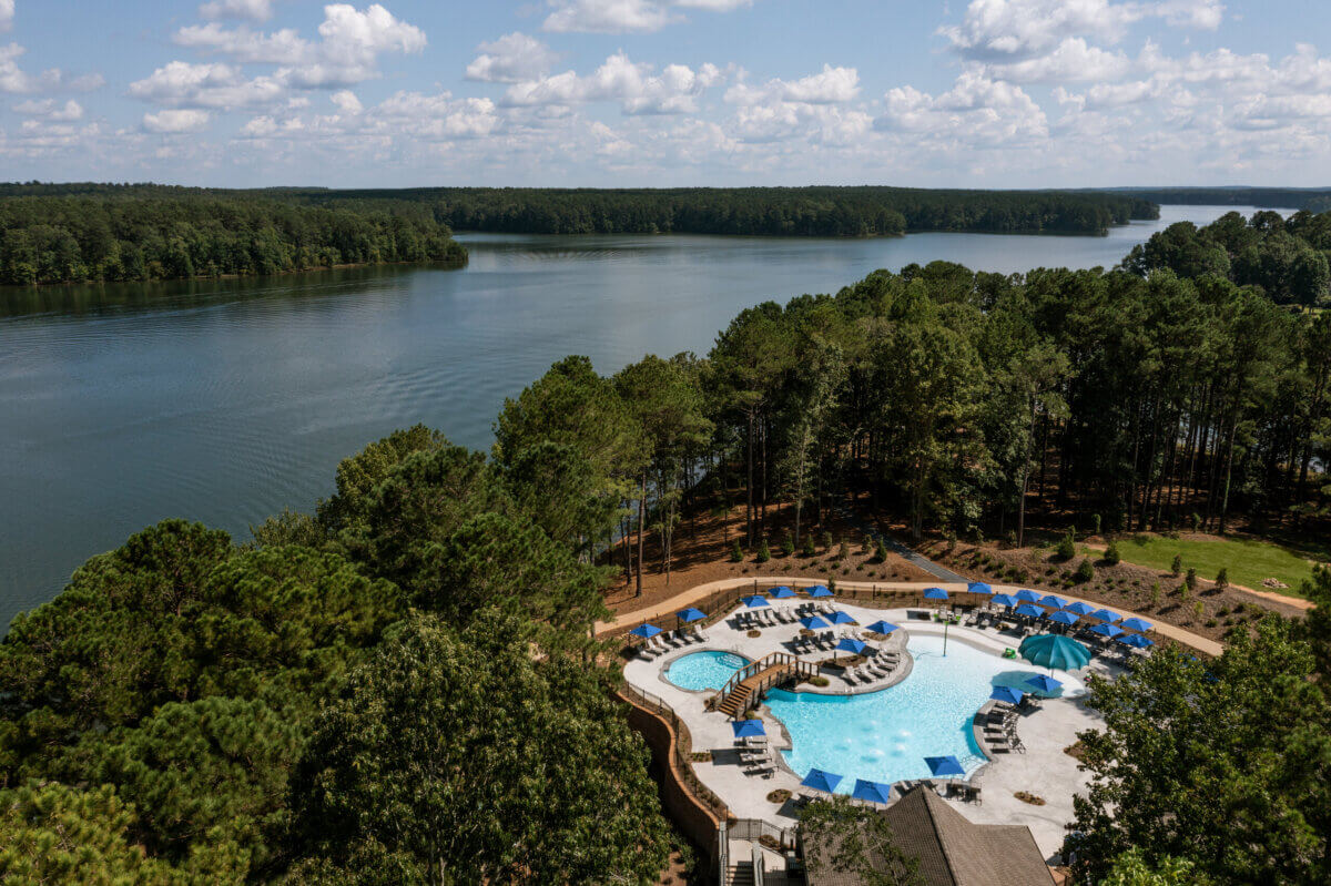 8 Luxurious, Family-Friendly Resorts in the South - 16 Aerial view of an outdoor swimming pool with blue umbrellas surrounded by trees near a large lake under a partly cloudy sky, offering the perfect retreat for Southern Families seeking family-friendly resorts.