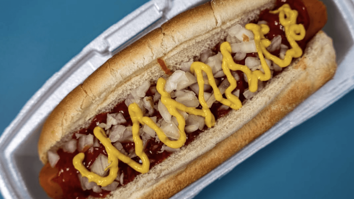 6 Louisville Hot Dogs Worth the Hype - 6 A hot dog in a bun with ketchup, chopped onions, and mustard in a zigzag pattern, served in a white foam container on a blue background.