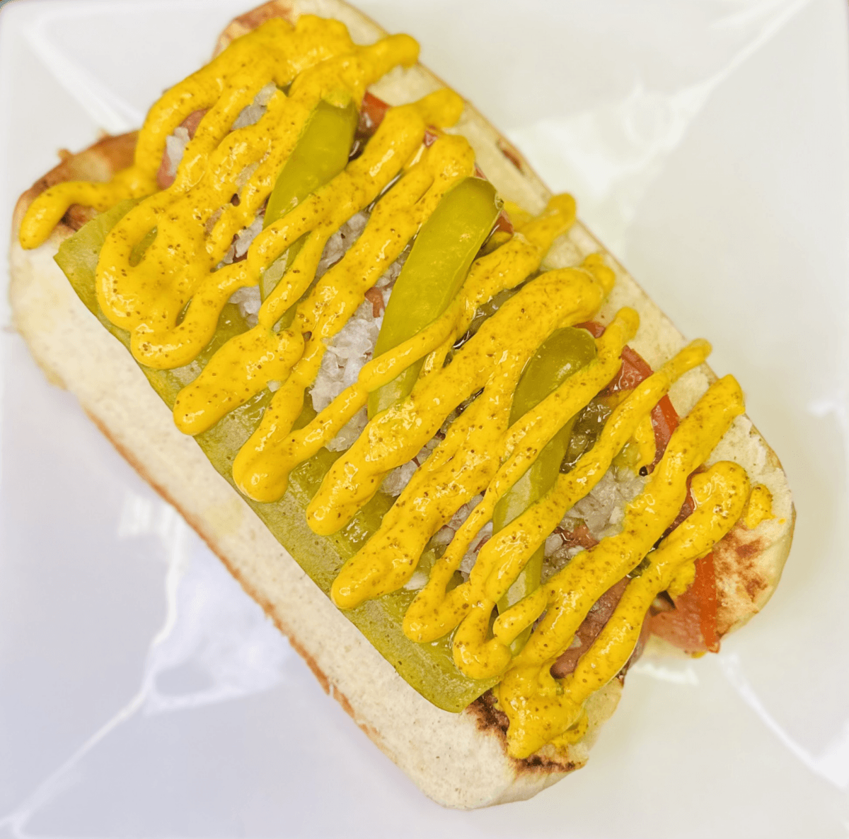 6 Louisville Hot Dogs Worth the Hype - 2 A hot dog in a bun topped with chopped onions, pickles, peppers, and yellow mustard, served on a white plate.
