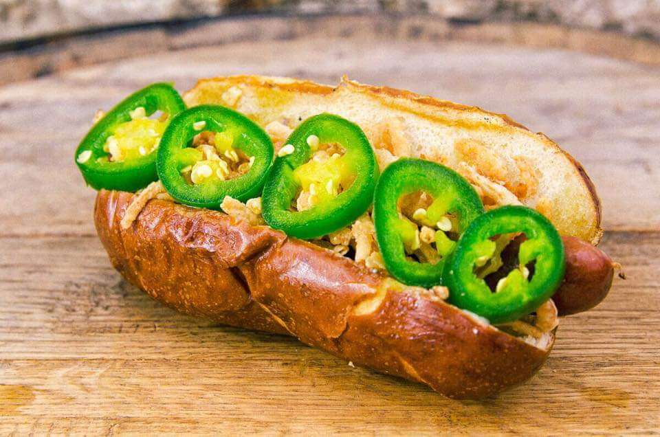 6 Louisville Hot Dogs Worth the Hype - 3 A hot dog in a pretzel bun topped with sliced jalapeño peppers and shredded cheese sits on a wooden surface.