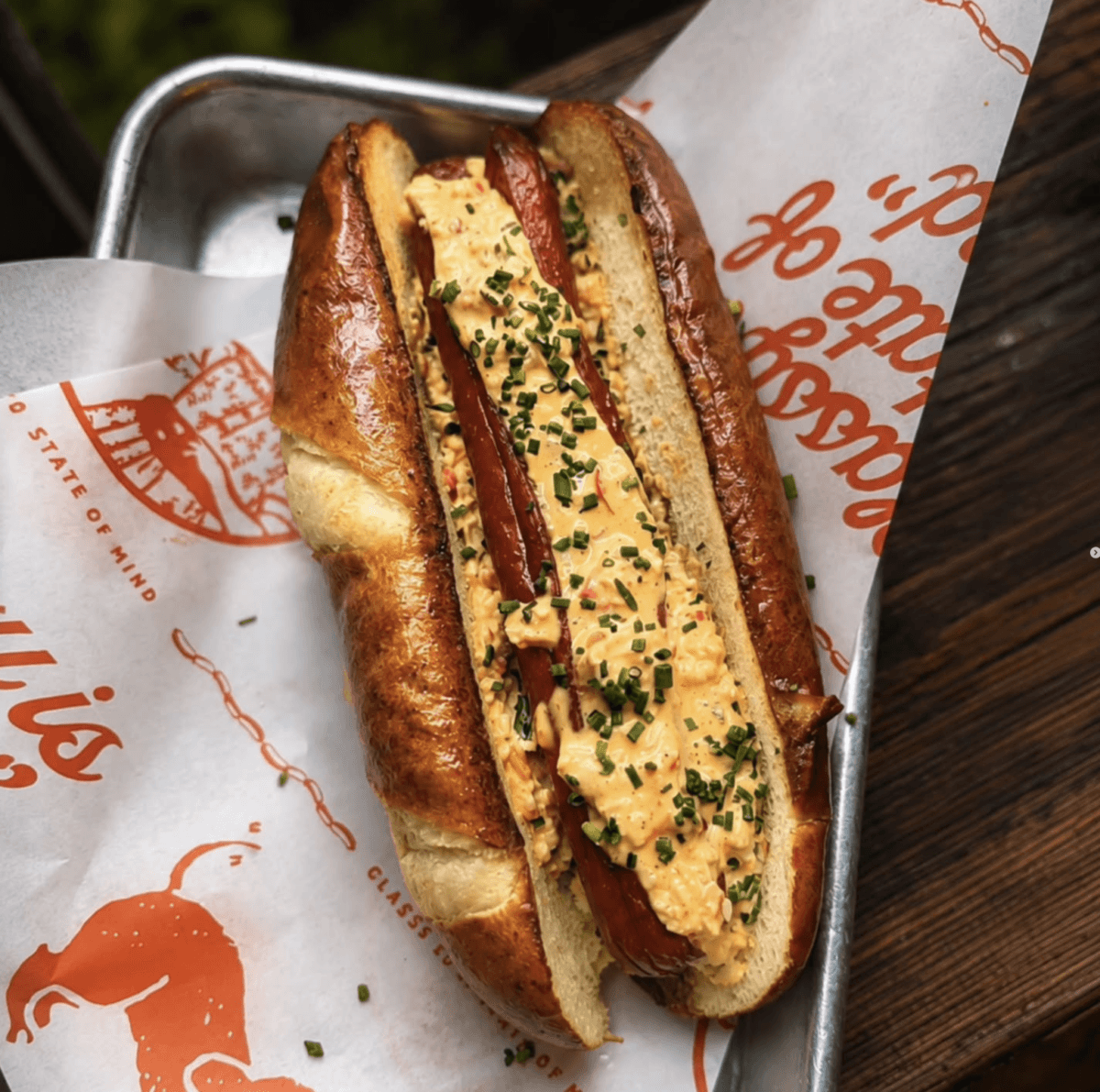 6 Louisville Hot Dogs Worth the Hype - 7 A grilled sausage in a split pretzel bun topped with a creamy pimento cheese spread and chopped chives, served on branded paper in a metal tray.