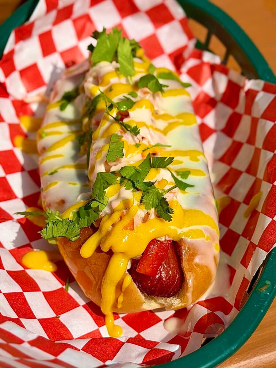 6 Louisville Hot Dogs Worth the Hype - 4 A grilled hot dog in a bun is topped with yellow mustard, creamy white sauce, and chopped cilantro, served in a green basket lined with red and white checkered paper.