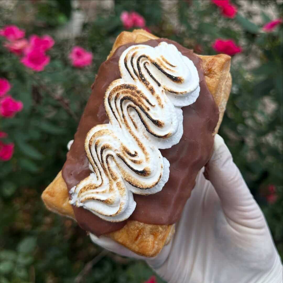 A gloved hand holds a pastry topped with chocolate glaze and swirls of toasted meringue, with pink flowers and greenery in the blurred background.
