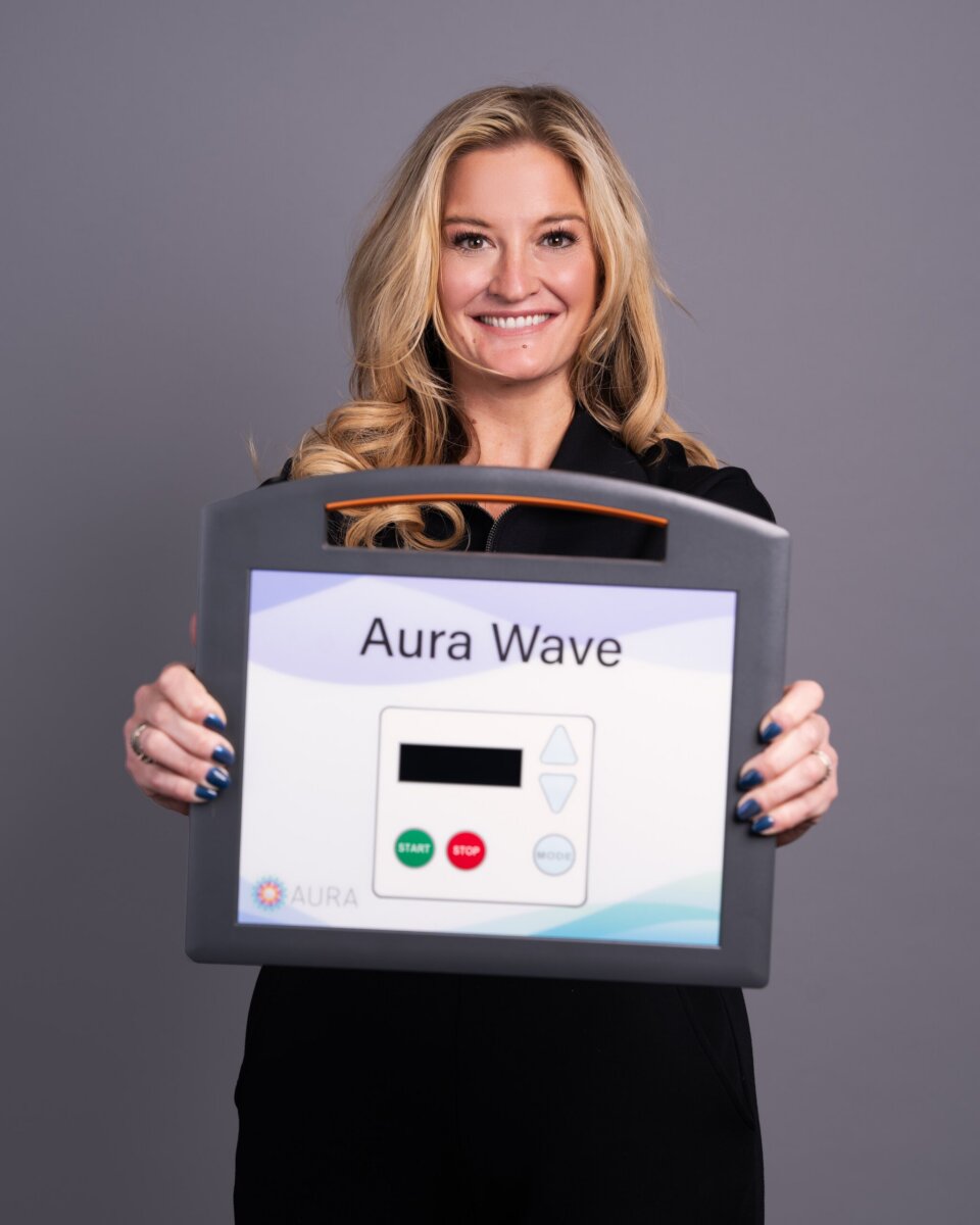 From TV Producer to Wellness CEO: Meet the Woman Revolutionizing Recovery - 2 A woman with long blonde hair holds a device labeled "Aura Wave," featuring a display screen and buttons, against a plain gray background—highlighting the rise of accessible wellness technology designed for women in tech.