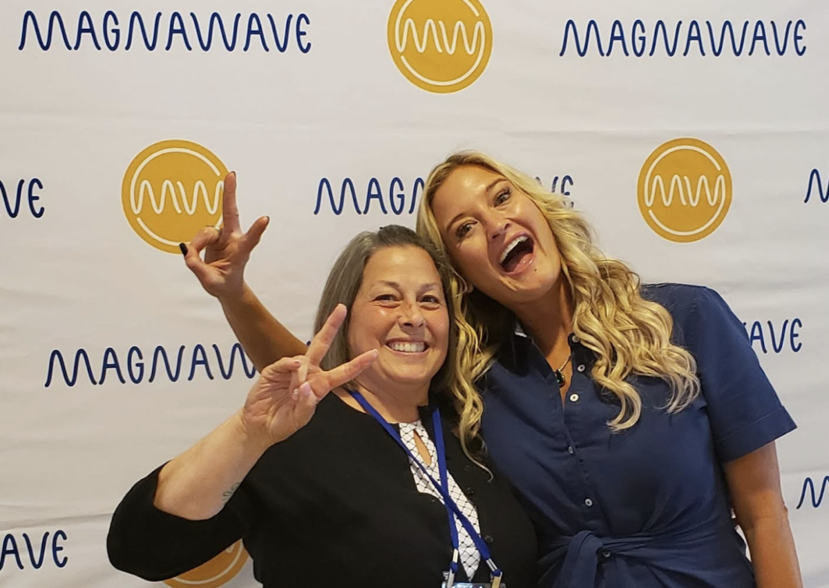 From TV Producer to Wellness CEO: Meet the Woman Revolutionizing Recovery - 3 Two women smile and pose with peace signs in front of a MagnaWave step-and-repeat banner, celebrating accessible wellness and the impact of women in tech.