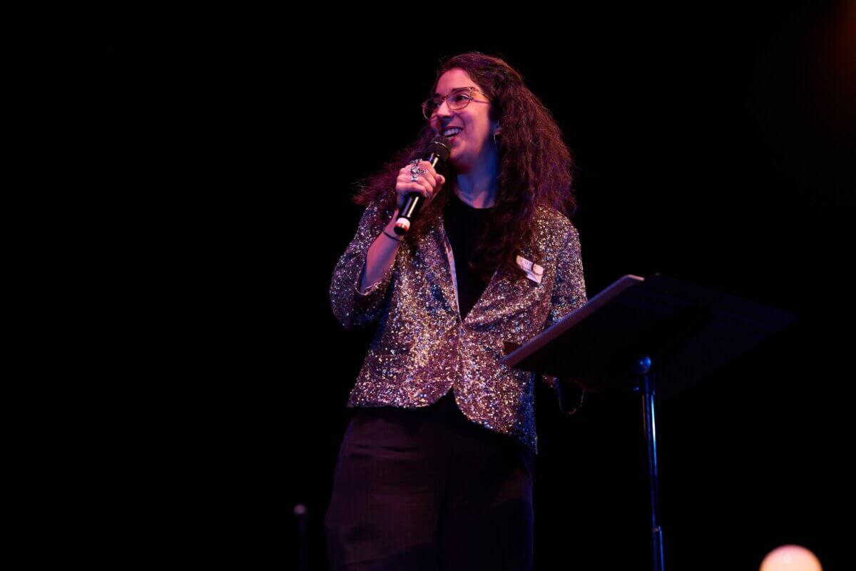 Meet the Creative Visionary Behind Actors Theatre of Louisville - 3 A person wearing glasses and a sparkly jacket speaks into a microphone on stage beside a music stand against a dark background.