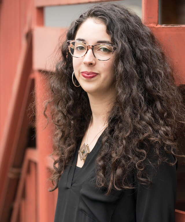 Meet the Creative Visionary Behind Actors Theatre of Louisville - 2 A woman with long curly hair, glasses, and hoop earrings stands in front of a red wooden structure, wearing a black top and geometric necklace.