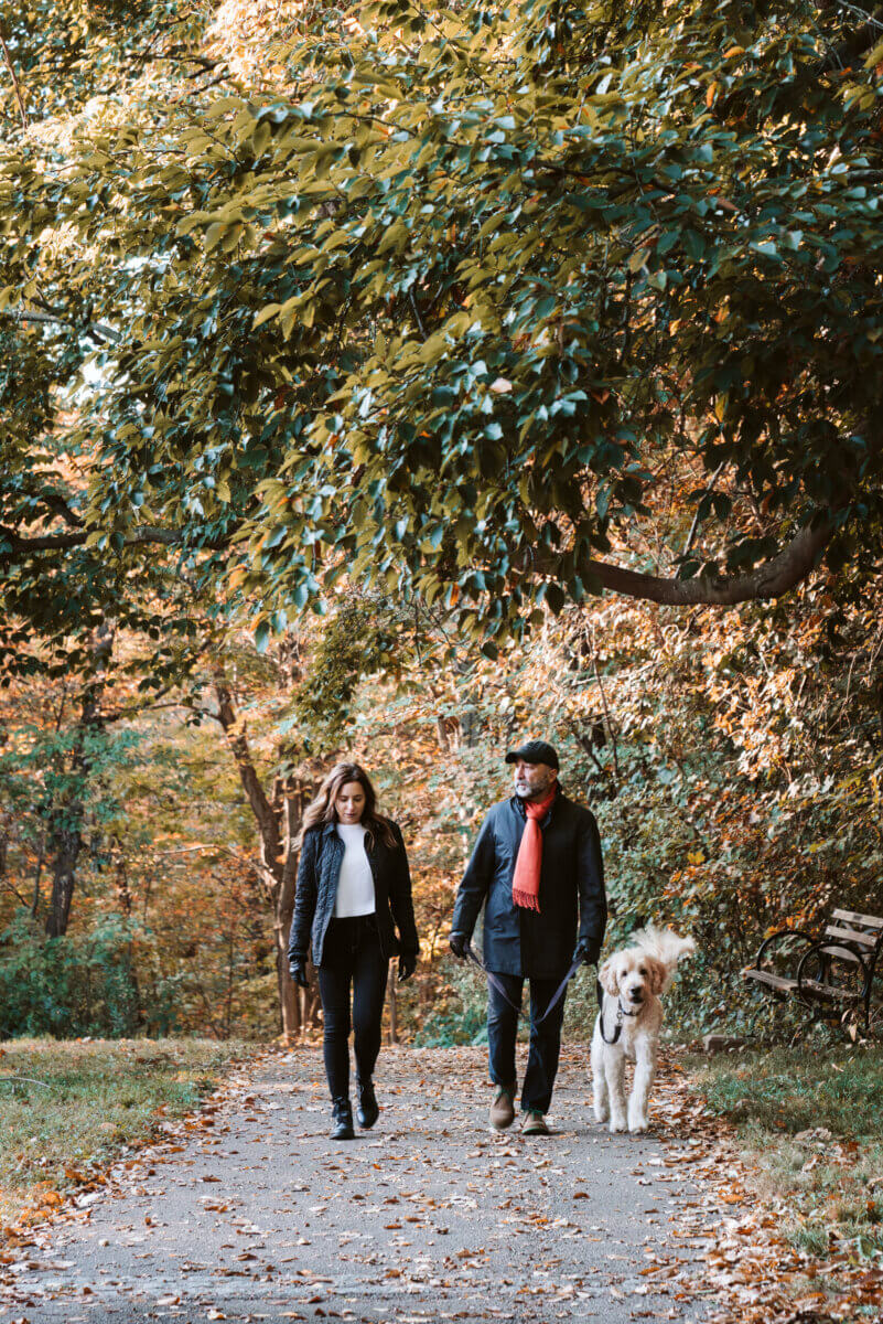 From Clinical Psychology to Cocktail Culture: Meet Heather Zamanian - 4 Two people wearing jackets and a red scarf walk a large, fluffy dog along a leaf-strewn path in a wooded area during autumn, drawing inspiration from the vibrant seasonβs colors.