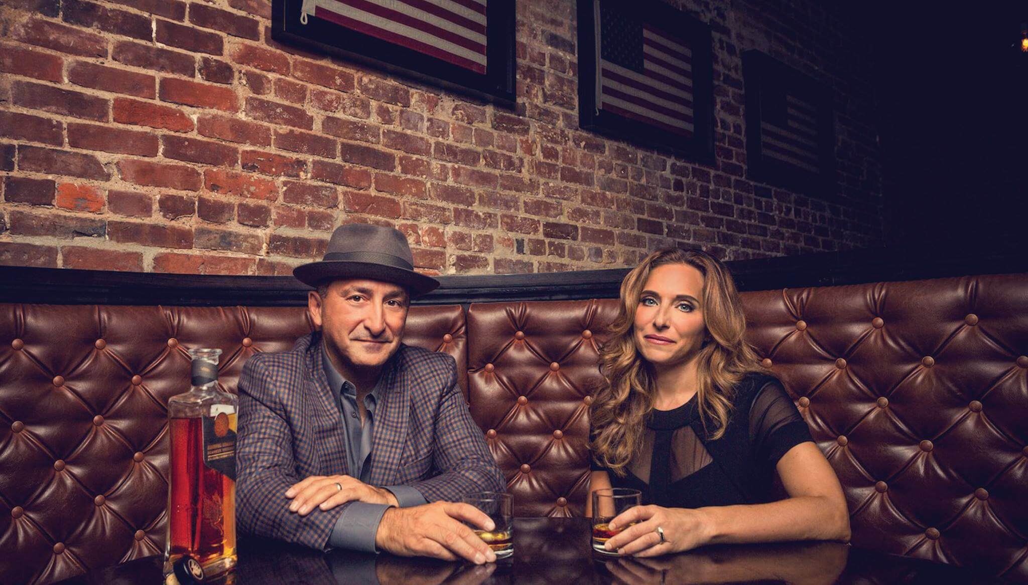 A man in a hat and a woman sit at a brown leather booth, drinks in hand, with a bottle of Rabbit Hole Distillery whiskey on the table and American flags decorating the brick wall behind them, adding inspiration to their setting.