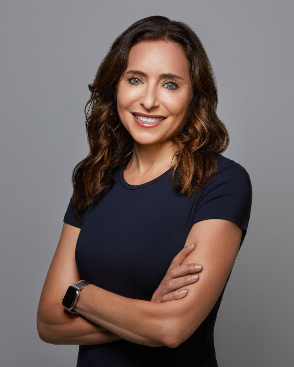 From Clinical Psychology to Cocktail Culture: Meet Heather Zamanian - 2 A woman with wavy brown hair, wearing a navy blue short-sleeve shirt and a smartwatch, stands with arms crossed and smiles at the camera against a plain gray background.