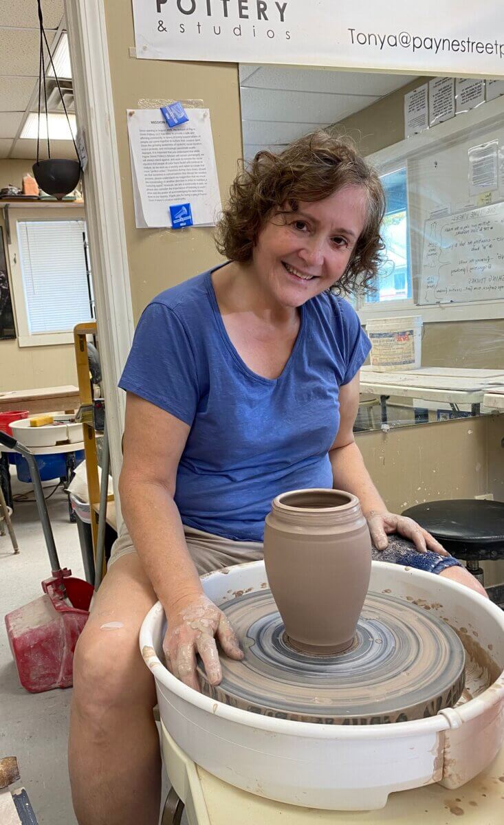 Meet the Louisville Artist Behind Payne Street Pottery - 2 A woman sits at a pottery wheel, shaping a lump of clay into a vase with her hands. She is smiling, wearing a blue shirt and shorts, inside a vibrant pottery studio that feels alive with the spirit of the pottery community.