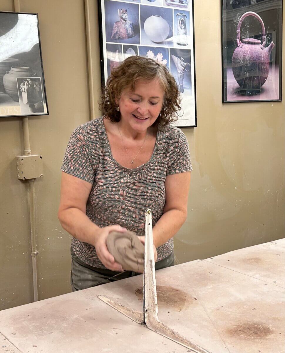 Meet the Louisville Artist Behind Payne Street Pottery - 3 A Louisville potter shapes a lump of clay on a table in a pottery studio, with framed ceramic art and photos hanging on the beige wall behind her, reflecting the creativity and community spirit found within her craft.