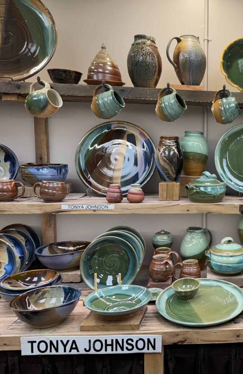 Meet the Louisville Artist Behind Payne Street Pottery - 6 Shelves display various handmade pottery pieces, including plates, bowls, mugs, and vases in earthy green and brown glazes. A sign reads "TONYA JOHNSON," celebrating a proud Louisville Potter whose clay creations enrich the local community.