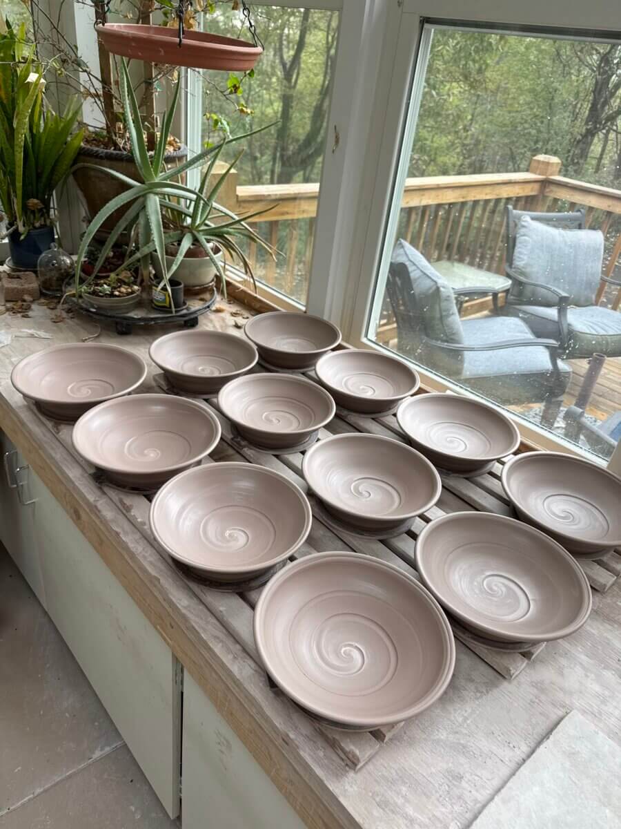 Meet the Louisville Artist Behind Payne Street Pottery - 4 Fourteen unglazed pottery bowls with spiral patterns drying on a wooden surface by a window, with plants and an outdoor deck visible in the background.