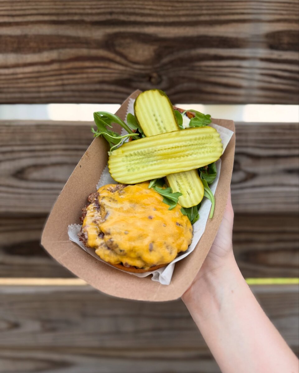 A hand holds a cardboard tray with a cheeseburger patty topped with melted cheese, leafy greens, and two pickle slices.