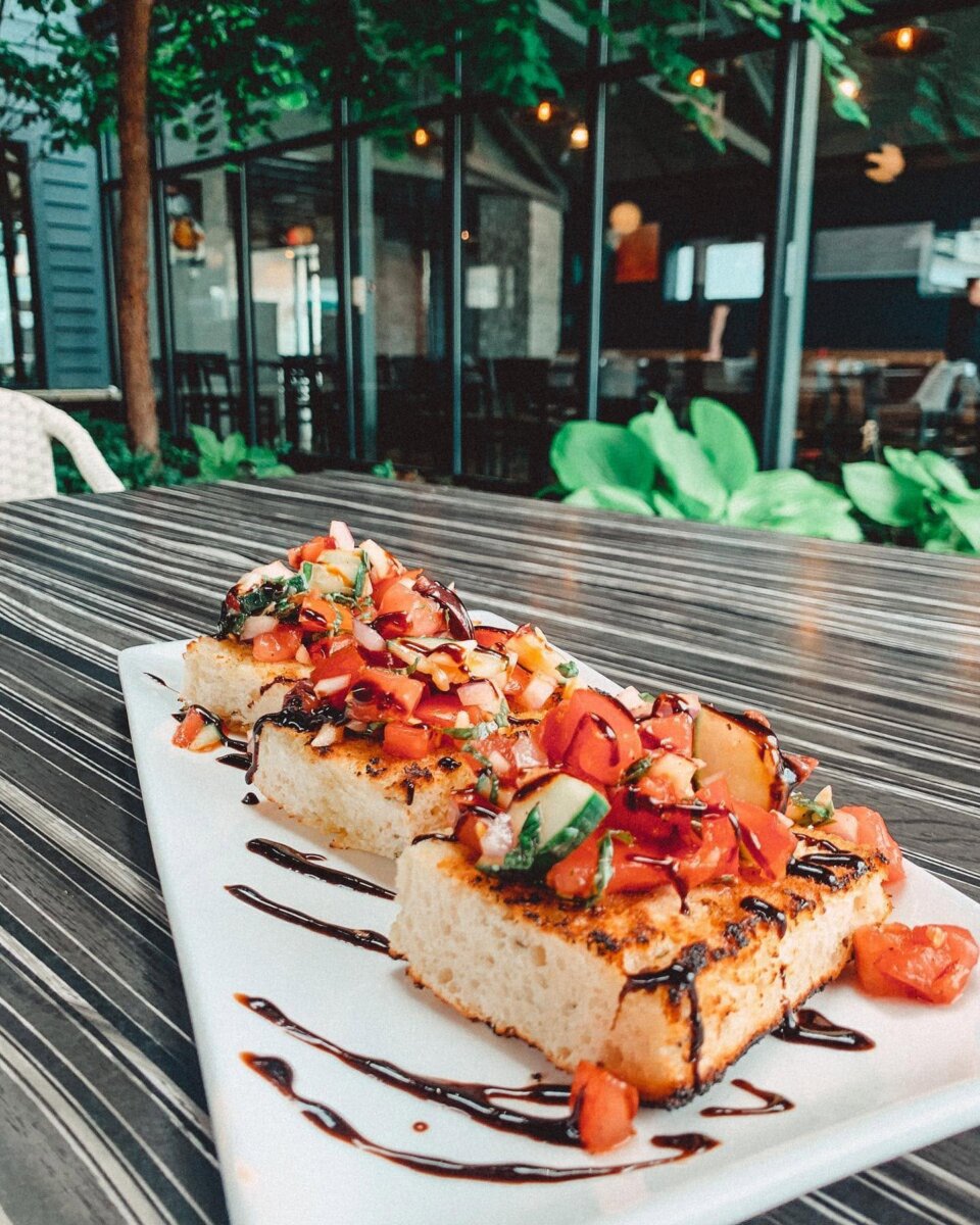 Three pieces of grilled bread topped with tomato bruschetta and balsamic glaze on a rectangular white plate, sitting on a wooden table at an outdoor restaurant in Germantown.