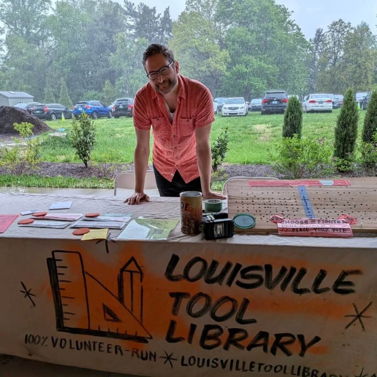 Shelf Help: Louisville's Tool Library Has You Covered - 2 A person stands behind a table with pamphlets and donation items at a Louisville Tool Library booth, offering shelf help to the community. The booth is set up outdoors, with cars parked in the background.