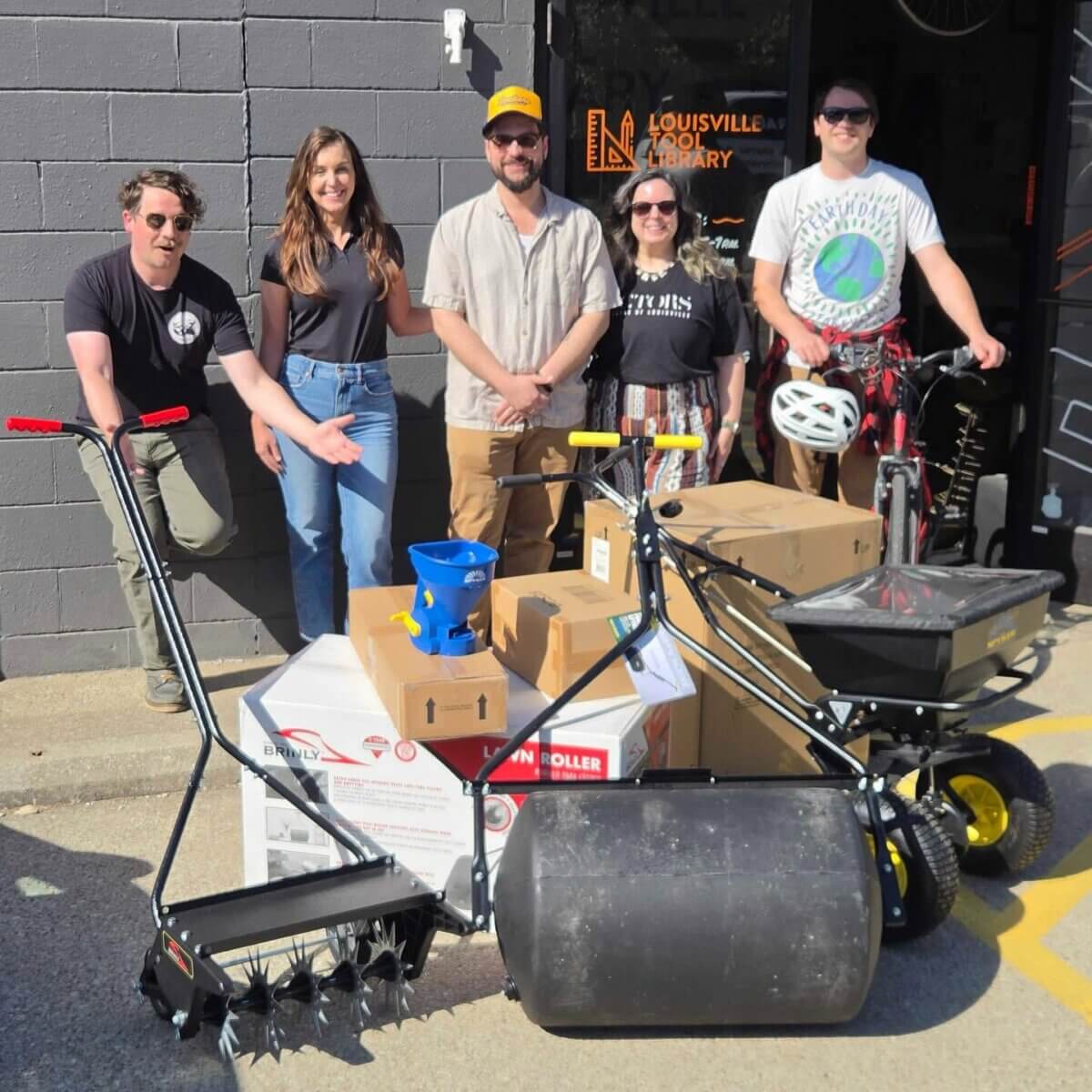 Shelf Help: Louisville's Tool Library Has You Covered - 5 Five people stand outside the Louisville Tool Library with various gardening tools, boxes, and a bicycle on a sunny day, ready to lend a hand through the Shelf Help program.