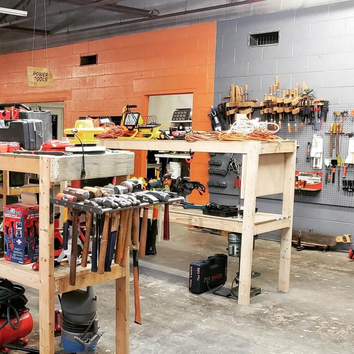 Shelf Help: Louisville's Tool Library Has You Covered - 4 Workshop with wooden workbenches, hand tools and power tools from the Louisville tool library neatly organized on shelves, pegboards, and tabletops; orange and gray painted walls in the background, offering true shelf help for every project.