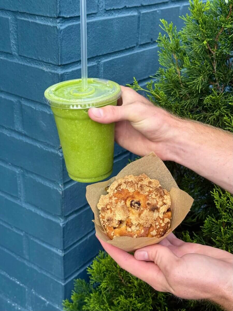 A person holds a green smoothie in a plastic cup and a muffin with crumb topping in front of a blue wall and green shrub, showcasing the fresh flavors often found at new restaurants enlivening the Louisville dining scene.