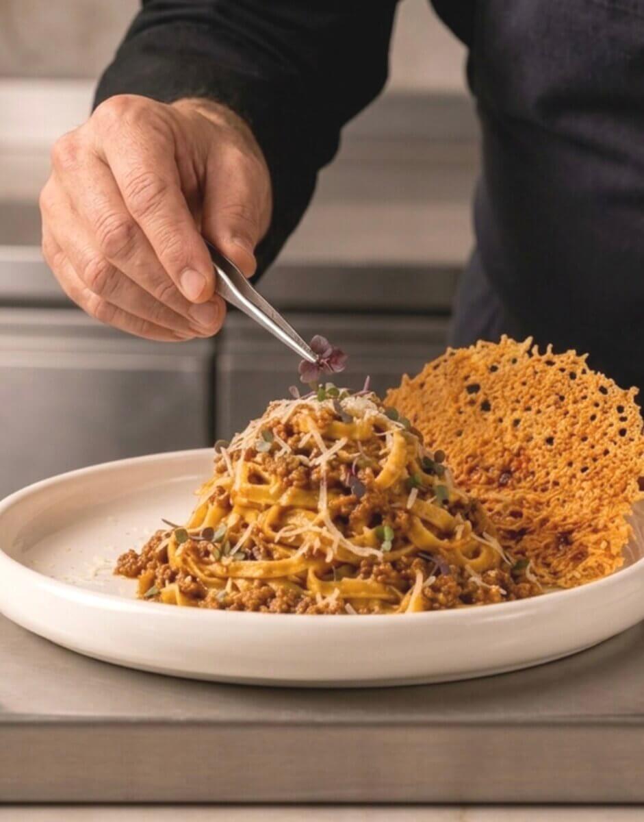 A person uses tweezers to place microgreens on a plate of pasta with meat sauce and cheese, next to a large crisp garnish—a touch of artistry reminiscent of the plating styles found in top Louisville restaurants.