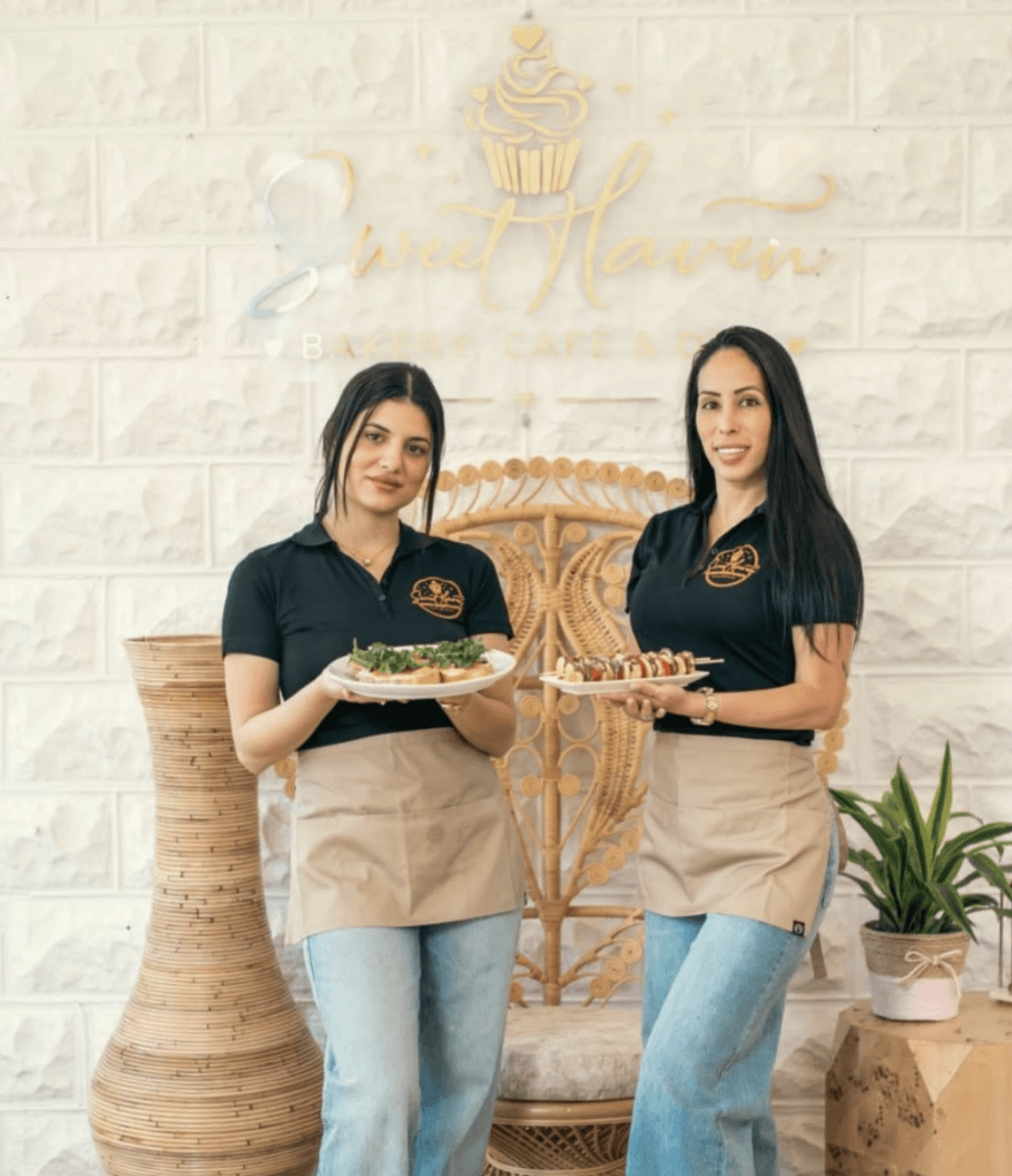 Two women in black shirts and beige aprons stand in front of a bakery sign, each holding plates of food. Showcasing the charm of Louisville dining, they stand by a peacock chair, woven lamp, and potted plant.