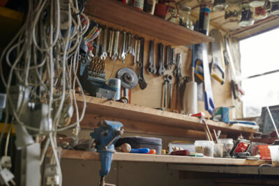 A cluttered workbench with tools, wires, jars, and various supplies; shelves above offer some shelf help with wrenches, cans, and equipment in a well-lit workshop.