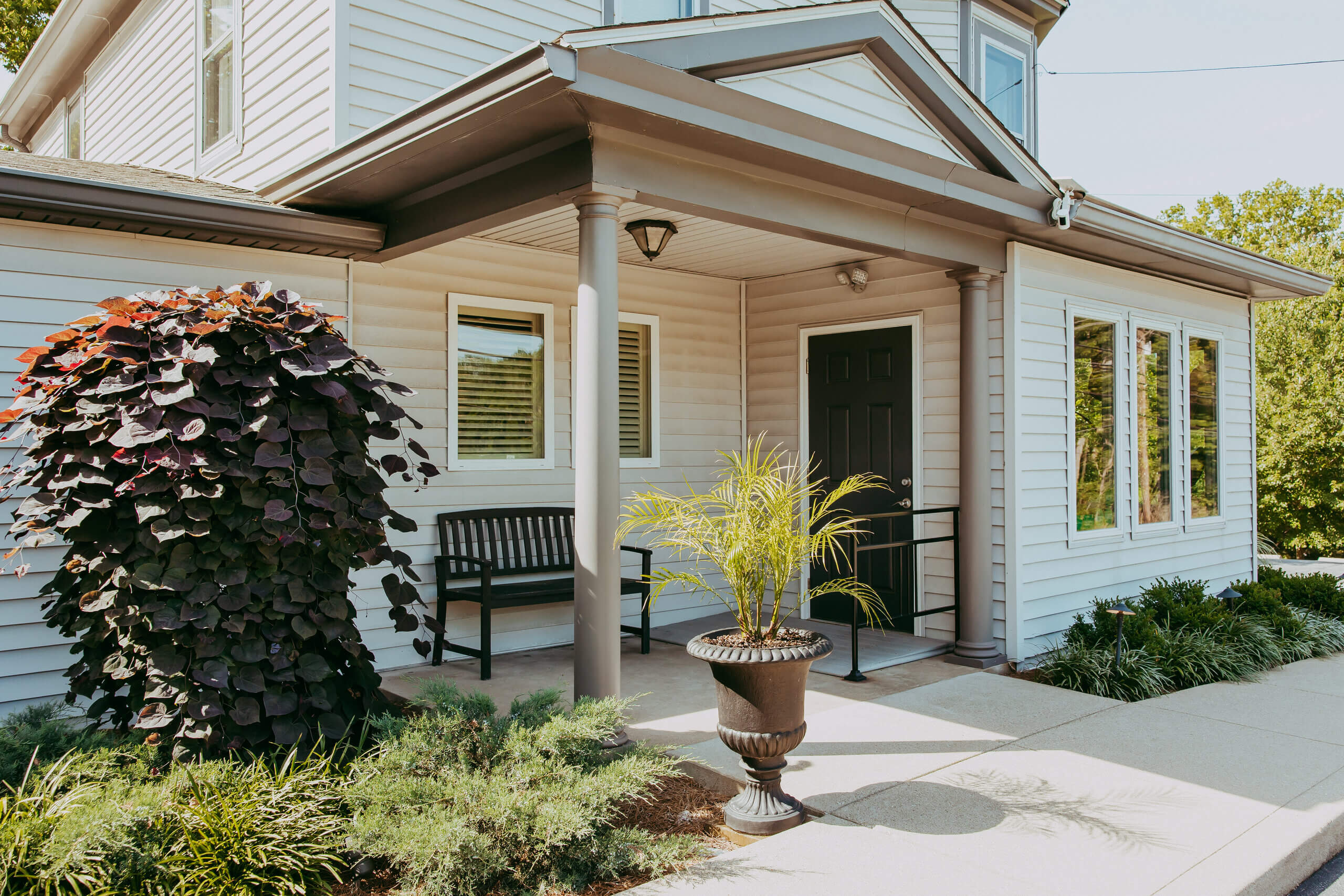 A house with white siding, a black front door, three large windows, a covered porch, a bench, and two potted plants flanking the entrance radiates charm—embracing the refined elegance of Vie Lavelle in its inviting design.