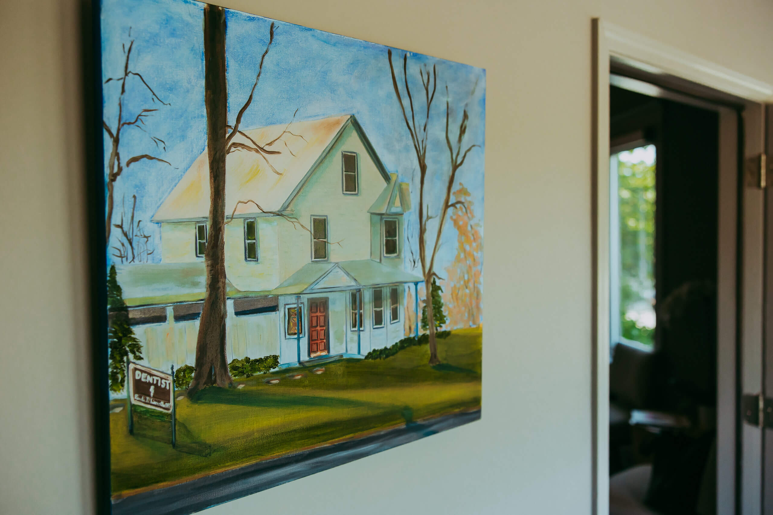 A painting of a white, two-story house with leafless trees and a “Dentist” sign hangs on a beige wall near a doorway, evoking the welcoming ambiance of Vie Lavelle Smile Design.