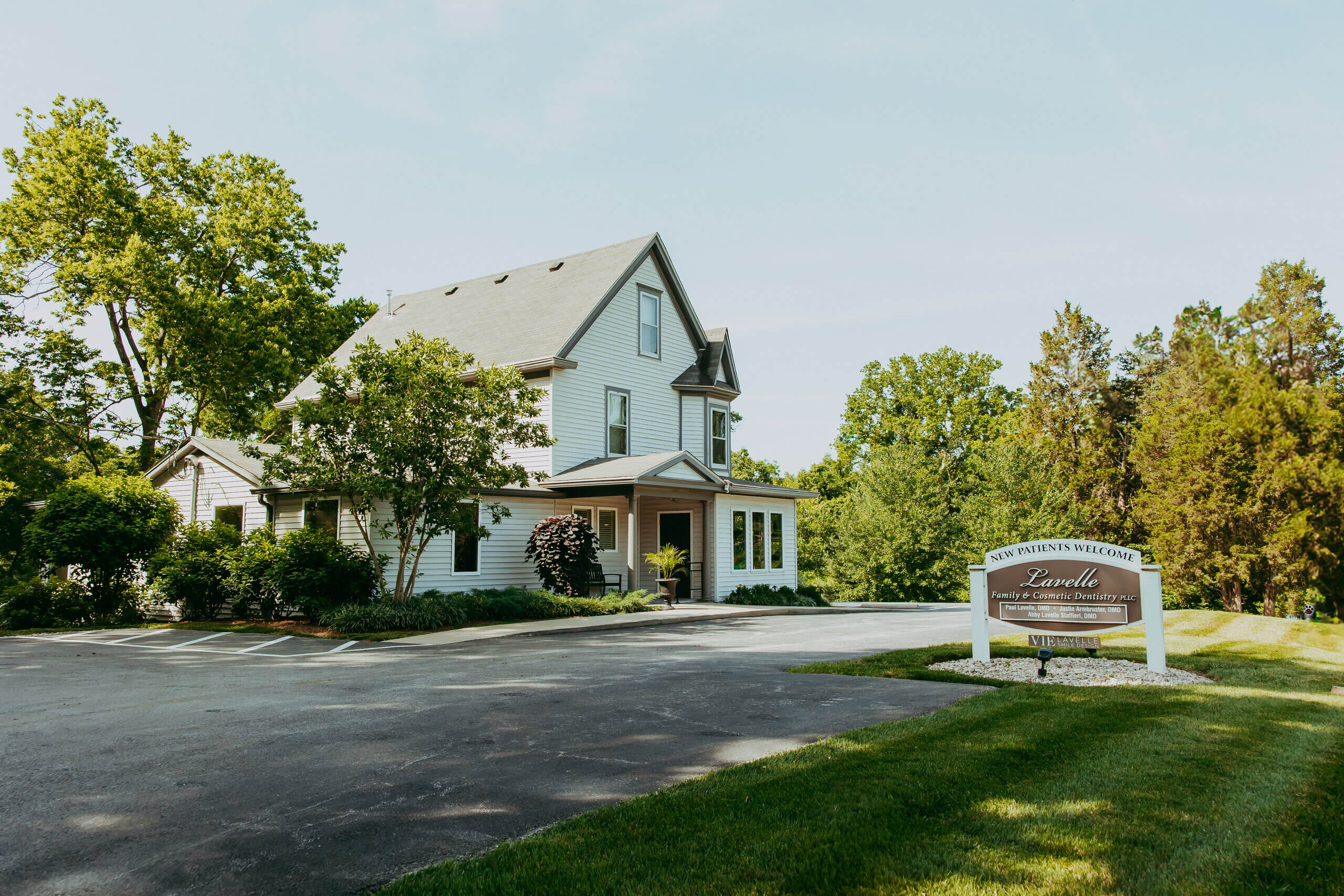 Two-story white house with a gray roof, surrounded by trees, with a sign in front reading “Vie Lavelle,” set beside a paved driveway and green lawn on a sunny day—a welcoming space where Smile Design begins.