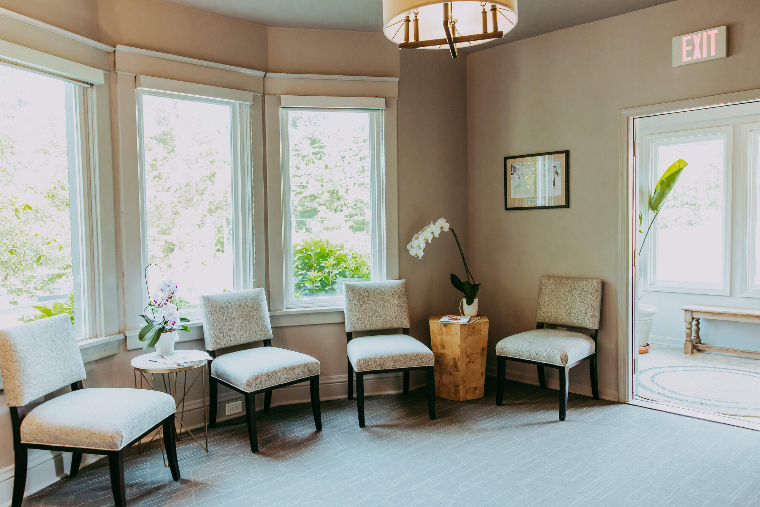 A waiting room with six beige chairs, a small table with a plant, large windows letting in natural light, and a framed picture on the wall creates an inviting atmosphere—perfect for welcoming clients to Vie Lavelle's Smile Design studio.