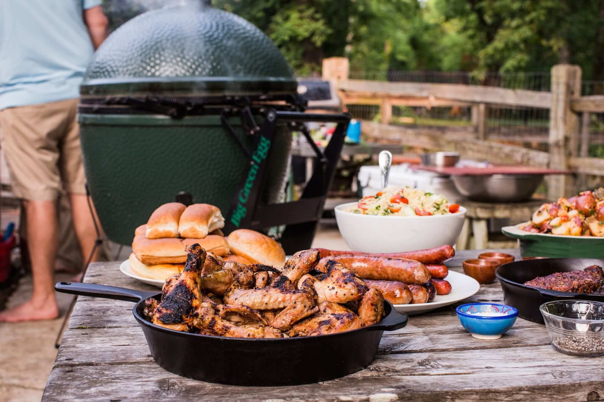 A picnic table set with grilled meats, sausages, burger buns, salad, and sides evokes the warm hospitality of the South, featuring a green grill and a person standing in the background.