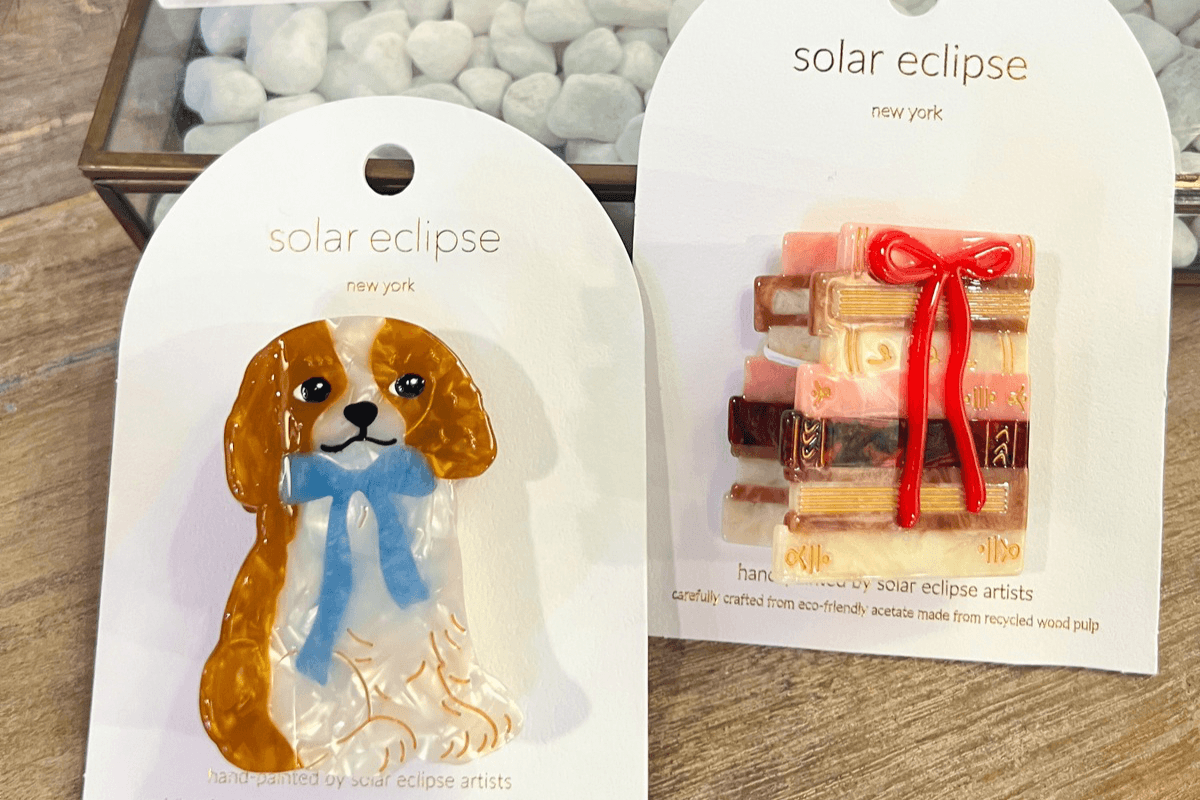 20 Memphis FINDS to See You Through the End of Summer - 9 Two decorative pins on display cards: one features a brown and white dog with a blue ribbon, the other shows a stack of books tied with a red bow. Both are labeled "solar eclipse new york.